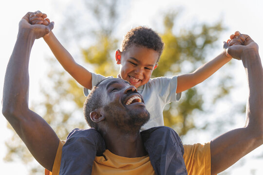 Loving African American father carrying his cheerful son on his shoulders while walking through a sunny grassy field, sharing joyful moments in nature.