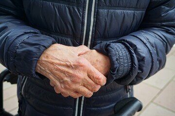 Übereinander gefaltete Hände von alter stehender Frau mit blauer Steppjacke auf Gehweg in Stadt bei Kälte und Frost am Nachmittag im Winter