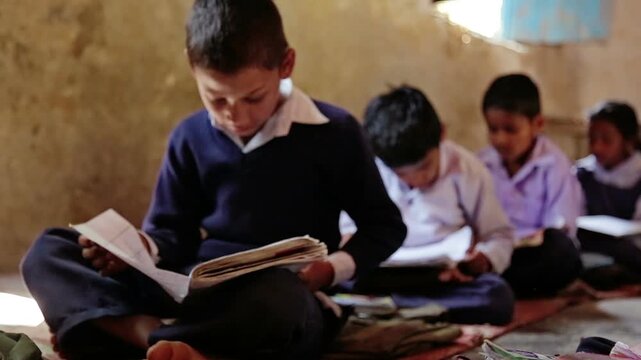 Raw footage of Indian village school boy reading textbook while sitting on classroom floor with other students inside rural government school in India.