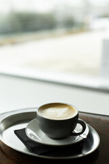 Hot cappuccino cup with latte art on a metal tray and dark napkin, sitting on a wooden table by a bright window