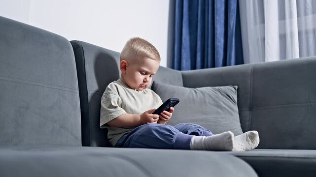 Toddler sitting on sofa with phone and looking at screen. Boy 2 years old playing with phone at home. Modern digitalization of children.