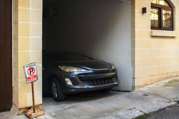 A dark car is poised to exit the doorway of a beige stone garage, set against a building with a window, right next to a warning sign reading 'No Parking, Garage in Use'