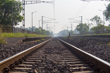 Low-angle view of a railway track stretching into the distance, surrounded by greenery and desho grass along the sides. The image captures perspective, depth, and a calm rural environment.