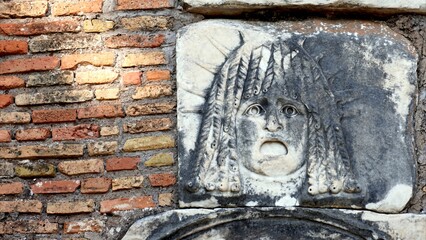 Ancient stone theatrical mask relief, weathered and textured against a worn Roman brick wall,...