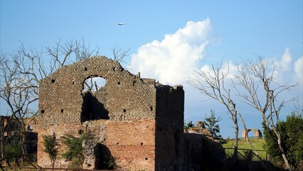 Ancient Roman ruins with crumbling stone and brick walls standing against a clear blue sky with an airplane flying overhead, symbolizing a unique intersection of history and modern life