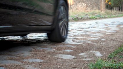 Car tire moving quickly along the historic Appian Way, a Roman street paved with large, time-worn cobblestones and surrounded by the serene atmosphere of ancient ruins