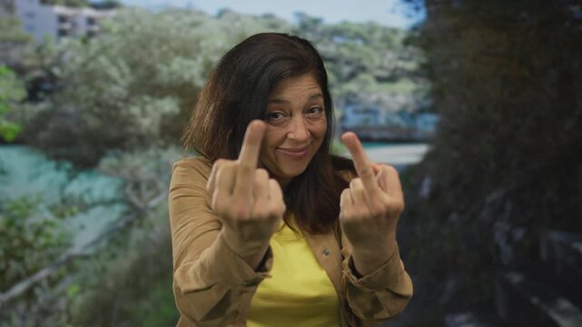 Woman flips middle finger in forest amid green trees while winking at camera, both middle fingers raised and in focus; defiance.