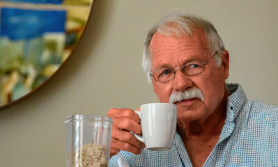 A senior man with a mustache and glasses holds a white coffee mug to his lips while looking thoughtfully at the camera.