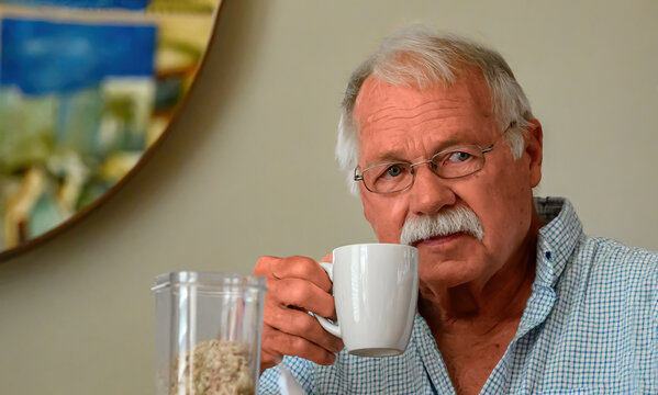 A senior man with a mustache and glasses holds a white coffee mug to his lips while looking thoughtfully at the camera.