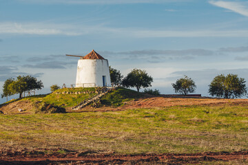 white windmill with a tiled roof sits peacefully on a green hill surrounded by scattered trees and farmland.