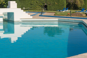 Bright sunny day by a shallow swimming pool featuring a stepped white structure and loungers on the grass.