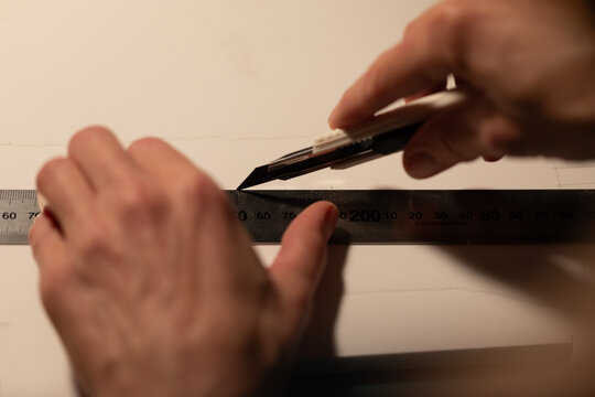 Over-the-shoulder view of hands cutting paper with a craft knife and ruler. Warm, focused light on a dark background highlights the meticulous process of creation and design.