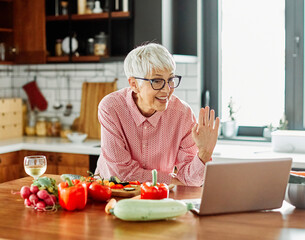 Portrait of happy senior mid aged mature woman prepering meal with fresh vegatebles and following internet instructions for a recipe on a laptop computer  or looking at video  or website app and drink