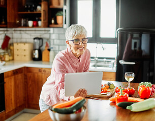 Portrait of happy senior mid aged mature woman prepering meal with fresh vegatebles and following internet instructions for a recipe on a tablet computer in kitchen
