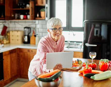 Portrait of happy senior mid aged mature woman prepering meal with fresh vegatebles and following internet instructions for a recipe on a tablet computer in kitchen - Powered by Adobe