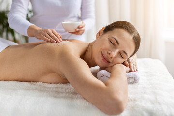 Young woman enjoying back massage while therapist applies exfoliating salt. Warm tones, gentle focus and soft texture highlight beauty care, skincare and deeply soothing spa treatment experience