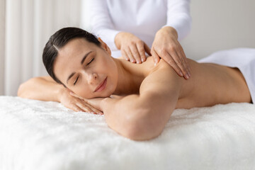 Young female smiling softly while receiving relaxing shoulder massage at spa. Warm light and gentle atmosphere emphasize comfort, beauty care, skincare and a joyful wellness lifestyle moment