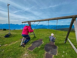A mother and her young child enjoy a playful moment on a wooden swing set situated on a lush green alpine meadow on Gerlitzen, Carinthia, with a spectacular panoramic view of the Austrian Alps.
