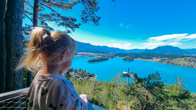 A young girl with blonde hair, seen from behind, stands at a scenic viewpoint and admires the breathtaking panoramic view of the turquoise Lake Faak and majestic Austrian Alps on beautiful sunny day