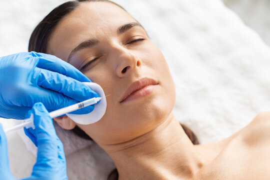Young woman receiving cheek area aesthetic injection using cotton pad support. Clean spa setting and precise gloved technique highlight cosmetic care, beauty enhancement and dermatology precision