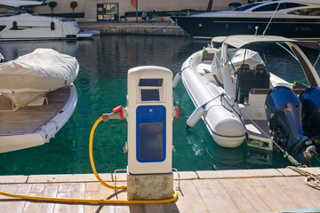Refueling station with a long yellow hose on a wooden dock of a luxury marina, with moored yachts and boats in bright turquoise water.