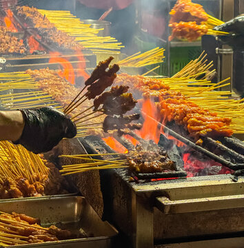 A street food vendor's gloved hand skillfully turns numerous skewers of delicious satay as they sizzle over glowing charcoal and open flames at the vibrant Lau Pa Sat hawker center in Singapore.