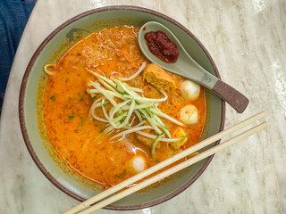 A delicious, steaming bowl of traditional Malaysian Laksa, a spicy coconut noodle soup with tofu puffs and fish balls, is served with sambal and chopsticks at restaurant in Melaka, ready to be eaten