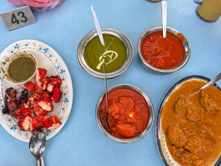 An overhead view of an authentic Indian meal in Melaka, Malaysia, featuring vibrant Tandoori chicken, a variety of rich curries, and savory chutneys served in traditional bowls on a blue table.