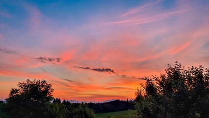 Expansive and dramatic sunset fills the sky with fiery palette of brilliant orange, soft pink, and deep blue, creating stunning, vibrant spectacle above dark silhouettes of trees and distant mountains