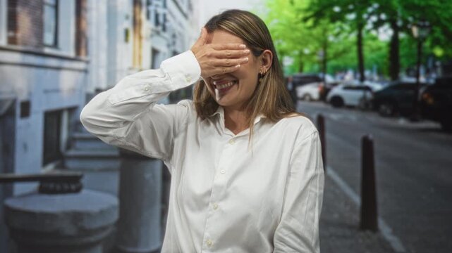 Woman covering her eyes with her hand while smiling and standing near parked cars on a city street in daylight, wearing a white button shirt; playful shyness.