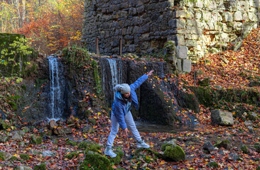 A person leans forward in a playful pose near cascading water and mossy rocks, surrounded by vivid autumn colors and rough stone ruins. The moment captures spontaneity and outdoor joy.