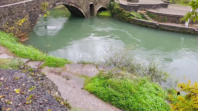 historic stone arch bridge spanning calm emerald river with mossy masonry, gentle eddies beneath arches, leafy banks and cobbled