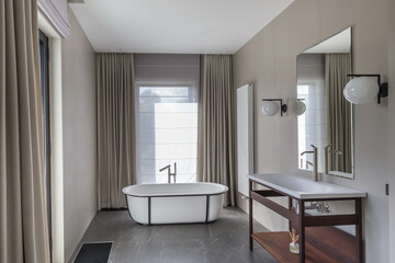 A spacious bathroom showcases a freestanding tub, stylish wall mirrors, and simple decor. Natural light filters through the large window, enhancing the relaxing atmosphere