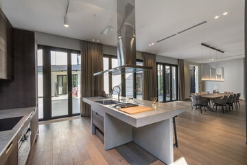 A spacious kitchen features a sleek island with a stove, modern appliances, and large windows that flood the area with sunlight. A dining area is visible in the background
