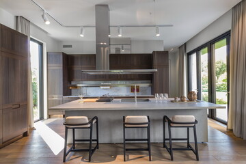 This stylish kitchen features a large island with three bar stools, modern cabinets, and windows that provide plenty of light. The open space showcases wood accents and is perfect for cooking