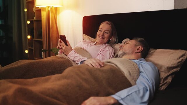 Middle-aged woman texting on phone next to sleeping husband, using dating app