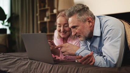 Happy middle-aged couple shopping together on laptop while lying in bed - Powered by Adobe