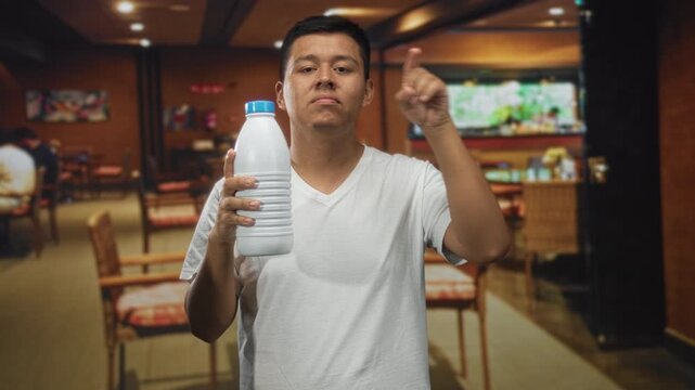 Man holding a white bottle with one hand and points finger forward in building restaurant interior; confidence product promotion.