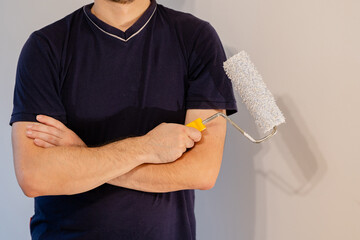 Mid-section portrait of a confident man in a dark blue t-shirt with his arms crossed, holding a paint roller. He looks prepared for a home renovation project.