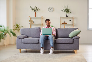 African american girl sitting at home on the couch in the living room with a laptop, close-up. Young woman in casual clothes online. Concept remote job, digital marketing, home work, search web