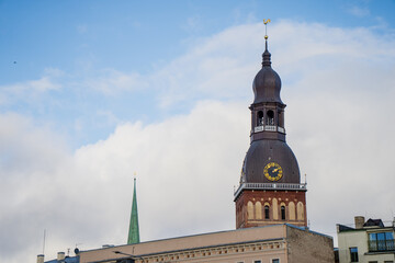 Riga church tower featuring rounded spire, ornate clock dial and layered brick facade emerging above surrounding buildings under open sky, representing prominent landmark in the Latvian capital