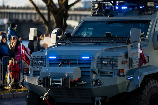 Heavily armored tactical vehicle displaying Latvian flags, protective panels, bright blue lights and large off-road tires moving near gathered spectators