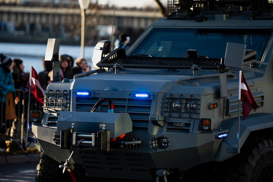 Military armored truck equipped with front winch, reinforced grille, Latvian flags and blue warning lights as it passes crowd lined behind barriers