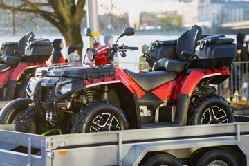 Utility ATV secured on transport trailer with visible storage cases, thick off-road wheels and front light cluster while pedestrians appear beside water in background © K