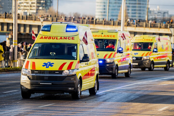 Yellow ambulances with blue emergency lights and Latvian flags moving in convoy along city roadway with medical symbols and red striping visible near bridge and crowd © K