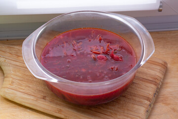 Top view of a homemade red borscht served in a transparent glass bowl. The vibrant red color of the broth contrasts with the wooden cutting board beneath it. Pieces of beetroot, cabbage, and vegetable