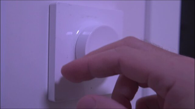 Close-up white round rotary dimmer switch on wall hand turning knob to increase and decrease light brightness then pressing to turn off modern lighting control