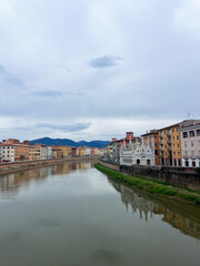 Tranquil river scene showcasing vibrant buildings in Pisa along the waterfront, with reflections in the water and mountains in the background, evoking a peaceful ambiance
