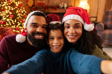 Portrait of happy African American family in Santa Claus hats celebrating Christmas and taking selfie, looking at camera and smiling