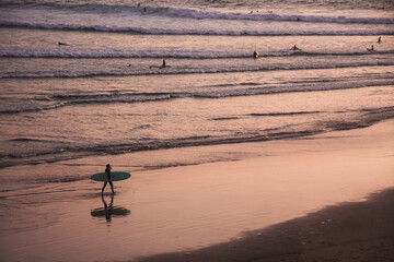 surfer walks along the beach at sunset carrying his surfboard, illuminated by soft purple and orange evening light